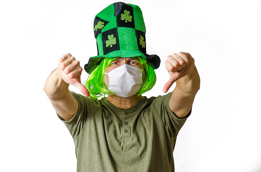 Portrait of man wearing a face mask, a green wig and hat for St. Patrick's day with clover leaves on white background, during covid-19 pandemic. He has two thumbs down.