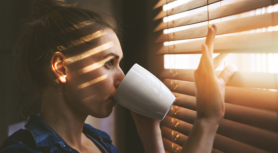 woman looking through window blinds into the sunlight and drinking coffee