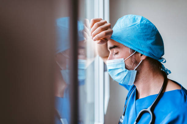 médico con máscara y uniforme apoyado en una ventana que parece agotado - doctor tired protective workwear sadness fotografías e imágenes de stock