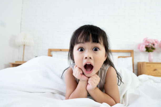 una niña asiática de 3 años que se despierta en la cama con una expresión sorprendente. - surprise child little girls shock fotografías e imágenes de stock