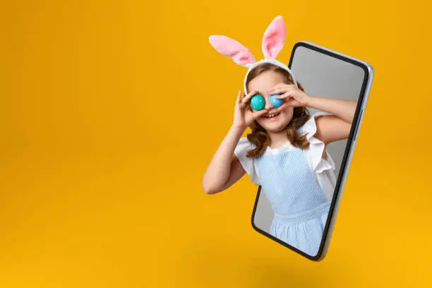 Photo of Happy easter. Smiling little girl in rabbit ears holds two eggs and looks out of a smartphone. The child is online in the mobile screen. Copy space.