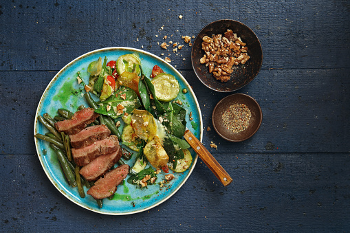 Grilled veal steak with green beans amandine. Spinach, grilled zucchini and walnut salad with potato chips. Flat lay top-down composition on dark blue background.