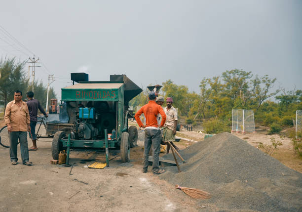 West Bengal, 01/25/2021: Rural development works at Sagar Island, during Gangasagar Mela. Group of manual labourers are working at a bitumen road construction site, with a butumen-asphalt mixer beside piles of stone-chip are seen on road.