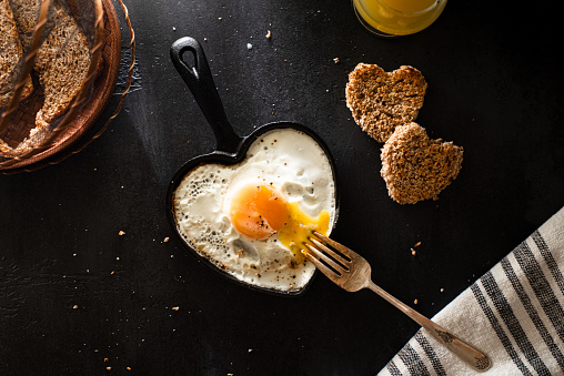 Flat lay of sunny side up egg in egg shaped cast iron skillet, orange juice, yellow berries and heart shaped bread on black background.