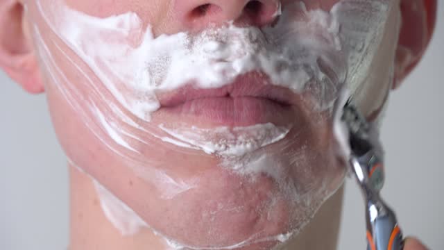 A young man shaves his face with a razor with foam or shaving gel close-up in the bathroom. Facial skin care, shaving of facial hair, stubble, mustache and beard.  Unshaven guy