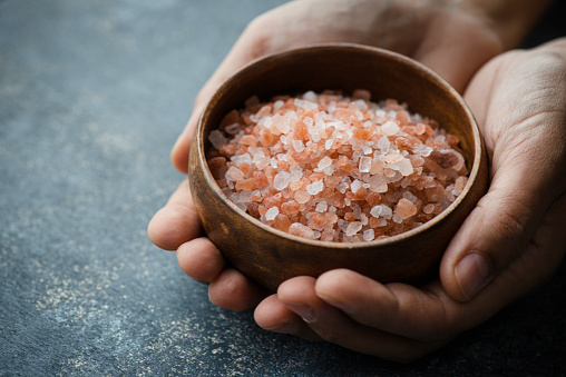 Hand is holding a bowl of Himalayan salt crystals to camera in front of dark blue background.