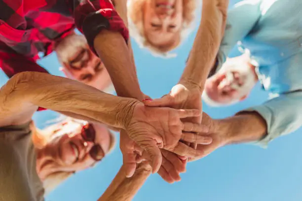 Low angle view of group of cheerful active senior people enjoying sunny summer day outdoors, gathered in circle, holding hands all together in the middle