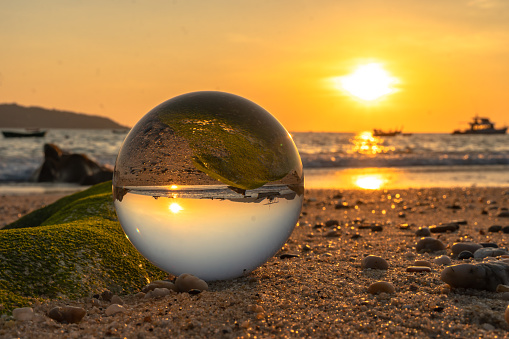 A crystal ball rests on the sandy pebbles of Kalim Beach. In the crystal ball, you will see an upside down view of the sunset.\