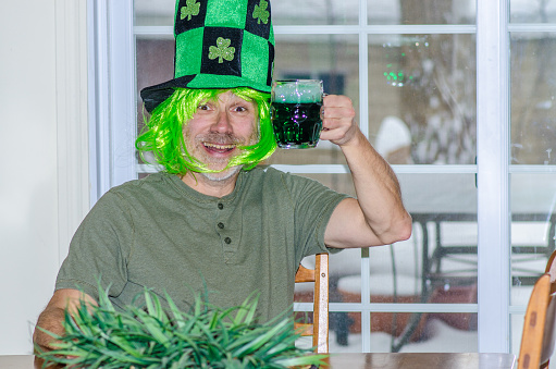 Portrait of man wearing green wig and hat for St. Patrick's day with clover leaves and having a green beer sit at his dining table