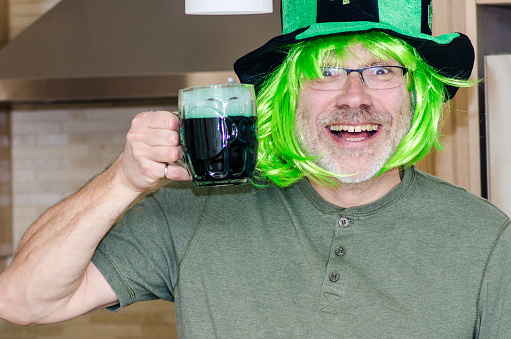 Portrait of man wearing green wig and hat for St. Patrick's day with clover leaves and having a green beer in his kitchen.