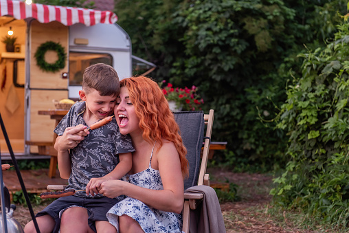 Happy family at picnic near the trailer track. Young mother and her son are sitting on a wooden deck chair, fooling around, eating sausages fried on a barbecue. Vacation outside the city, fun holidays