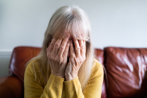 Color image depicting a sad, depressed-looking senior woman in her 70s, and of Caucasian ethnicity, sitting with her head in her hand at home. Room for copy space.