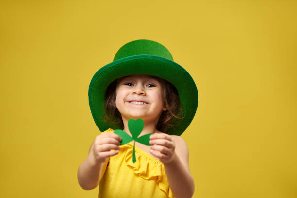 Pretty little girl holds a clover leaf in hands and shows it to camera. Saint Patrick's Day concept on yellow background. stock photo