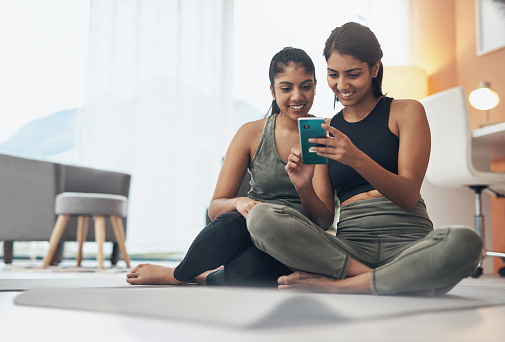Shot of two young women looking at something on a cellphone while sitting at home in exercise clothes