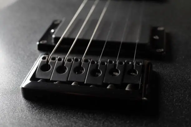 A close-up rusty bridge serves to secure the strings to an electric guitar. Black electric guitar in the dark. Selective focus.