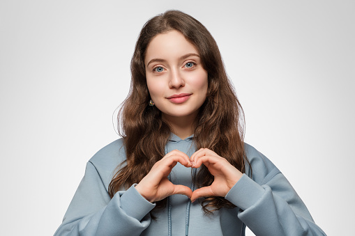 Young lady in a hoodie shows a heart with her hands. Long hair and blue eyes.