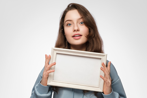 Young woman with an empty picture frame horizontally in her hand on a light blue background. The frame has a space for your text or photo. Light blue hoodie, blue eyes long hair.