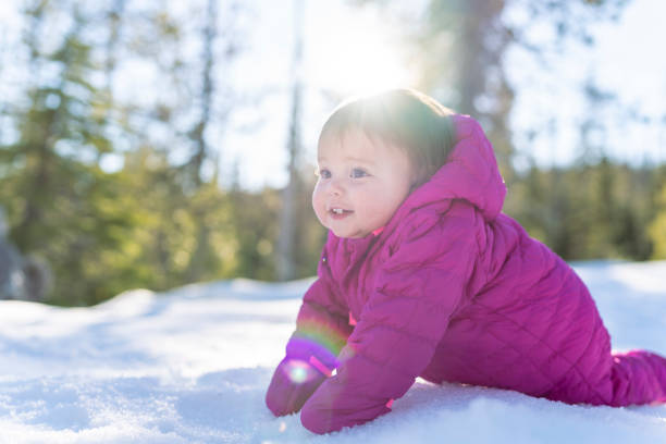 An adorable one year old baby girl wearing a pink snowsuit smiles while crawling in the snow for the first time.