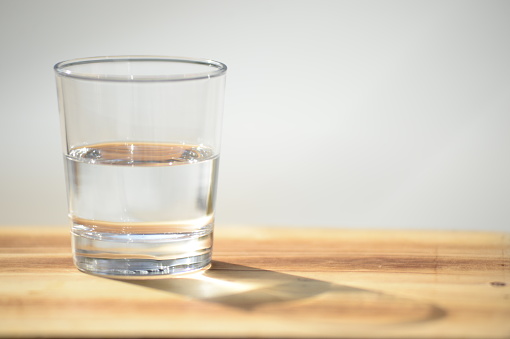 Half full or half empty glass of crystal clear water on wooden surface with bright blurred out background and copyspace with optimistic feeling, left justified