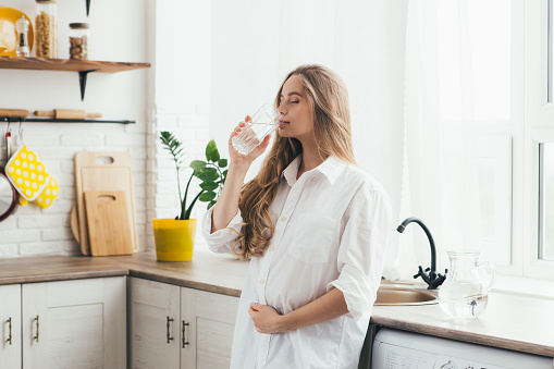 Young cute pregnant girl drinking clean water in the kitchen. The concept of a healthy lifestyle. Toning.