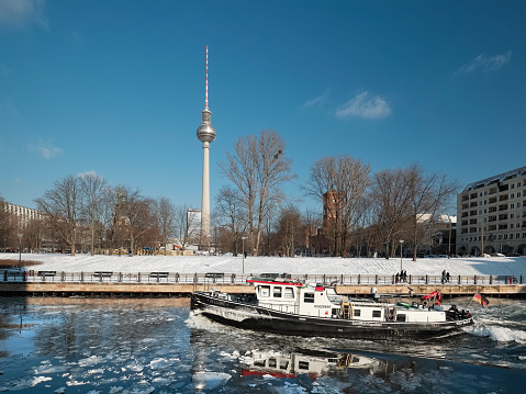 Berlin, Germany - February 10, 2021: : Berlin riverside in winter. Boat crashes ice on Spree river. Panoramic image with city skyline, Alexanderplatz television tower, blue sky and white snow.