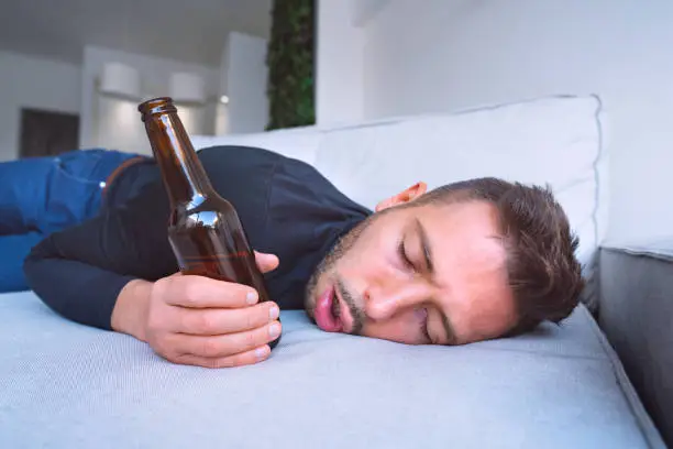 Photo of Drunk young man with open mouth and bottle of beer sleeping on the sofa at home.