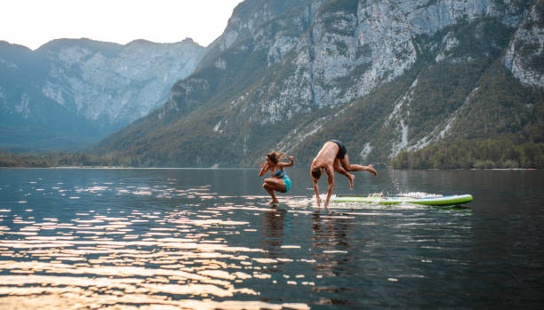 vacationing couple jumping off paddleboard into lake bohinj - lake bohinj imagens e fotografias de stock