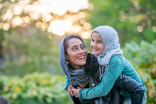 A beautiful Muslim mother is giving her daughter a piggy back while outdoors at the park. They are both happy and smiling.