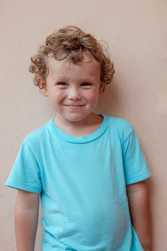 Portrait of a charming curly blond boy in a blue T-shirt. Child looks into the camera and smiles slyly.