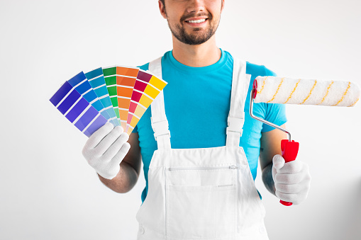 Crop man in uniform smiling and showing color palettes and paint roller during renovation works against white wall