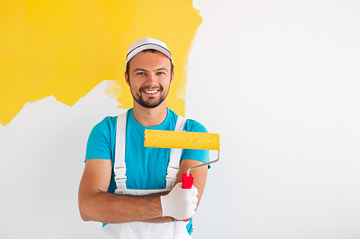Confident professional male painter in workwear with paint roller looking at camera and smiling friendly while standing against partially painted in yellow color wall