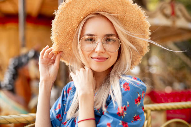 Charming young woman in hat near carousel Attractive young blonde female in trendy glasses and colorful wear touching straw hat and looking at camera while standing against blurred carousel in amusement park straw hat photos stock pictures, royalty-free photos & images