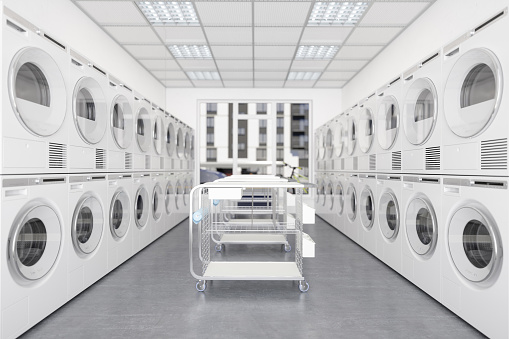 White Laundry Machines And Dryers In A Row In Laundromat With Wheeled Laundry Baskets.