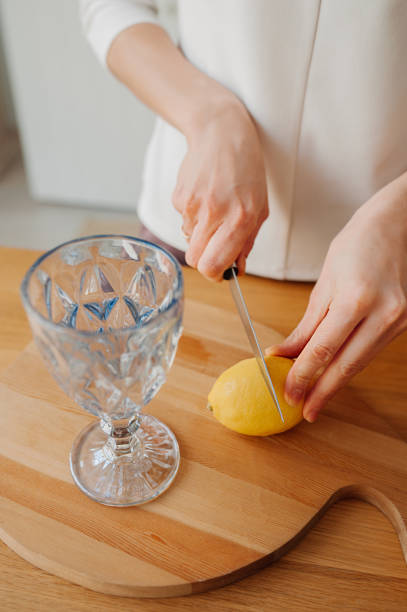 istock step-by-step instructions for preparing lemon water. Step 1. woman with no face slices lemon near glass of water in light kitchen. vertical wellness content, selective focus 1302566100