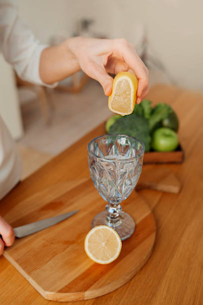 istock step-by-step instructions for preparing lemon water. Step 5. woman with no face squeezes the juice from lemon into glass in light kitchen. vertical wellness content, selective focus 1302566106