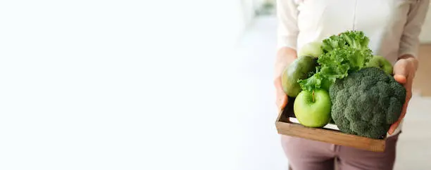 Photo of long format banner woman no face holds wooden eco storage box with green detox foods: apple, broccoli, lettuce, avocado. healthy food delivery, vertical food content, selective focus