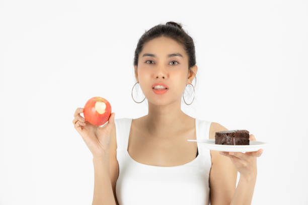 istock Upset stressed Asian woman choosing between apple fruit or unhealthy cake on hands over white isolated background. Healthy lifestyle and dieting concept. 1302570846