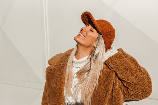 Portrait of a beautiful young woman with long blond hair wearing a fake fur brown color coat and fashionable brown color cap in the city