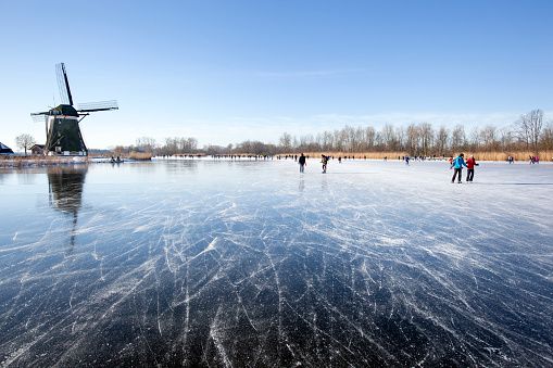 Zevenhuizen, Netherlands - February 13, 2021: Dutch winter landscape with Ice skaters on the Rottemeren near a windmill