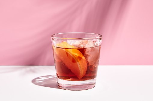 Negroni cocktail over pink background. Drink in rox glass in daylight with palm leaf hard shadow. Summer, tropical, fresh drink concept.
