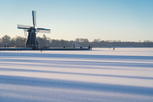 People ice skating during a tranquil winter sunrise near a windmill.