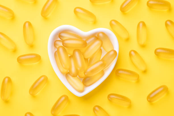 vitamina d y omega 3 cápsulas de aceite de pescado suplemento en una placa en forma de corazón sobre fondo amarillo. concepto de atención médica. vista superior - ácido grasos fotografías e imágenes de stock