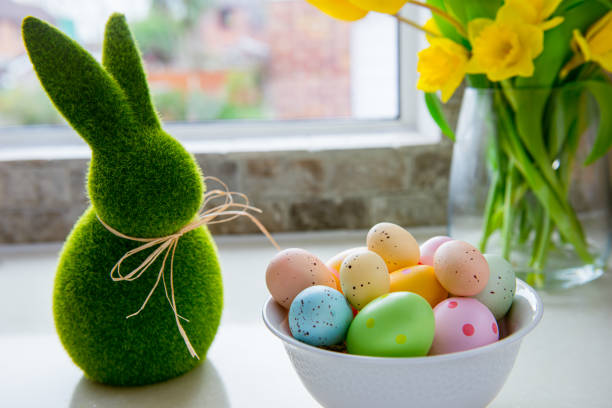 tazón blanco con huevos de pascua de colores, ramo de tulipanes amarillos y flores de narcisos y conejo conejito verde en la mesa de la cocina blanca cerca de la ventana. tarjeta de primavera ester festiva. enfoque selectivo. copiar espacio. - daffodil easter egg hunt easter easter egg fotografías e imágenes de stock