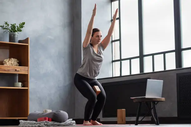 Photo of Caucasian woman doing Utkatasana, Chair Pose using block at home