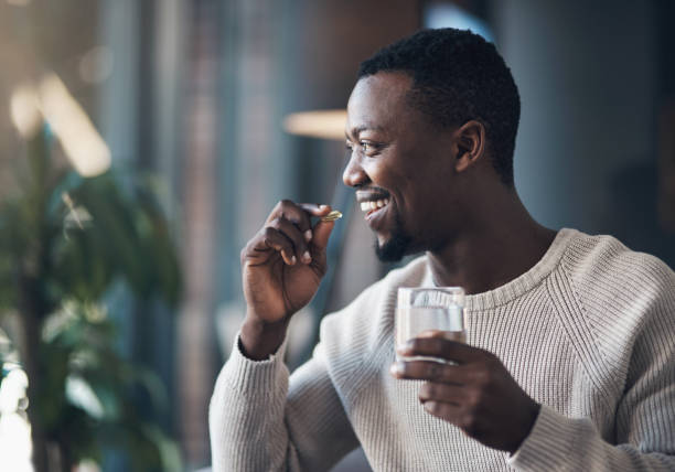 I never skip my vitamins Cropped shot of a handsome young man sitting alone and taking his medication with water in his living room relief emotion stock pictures, royalty-free photos & images