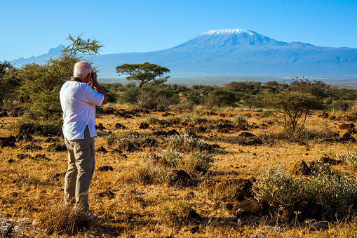 Kenya Prairies, Africa. Bushes and acacias at the foot of the greatest African volcano Kilimanjaro. Middle-aged gray-haired tourist in a white T-shirt photographs a magnificent landscape