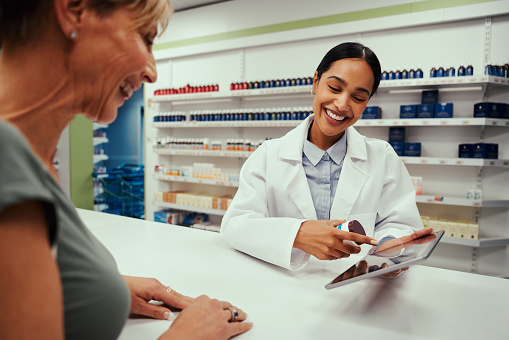 Cheerful young woman working in pharmacy showing screen to female customer on digital tablet