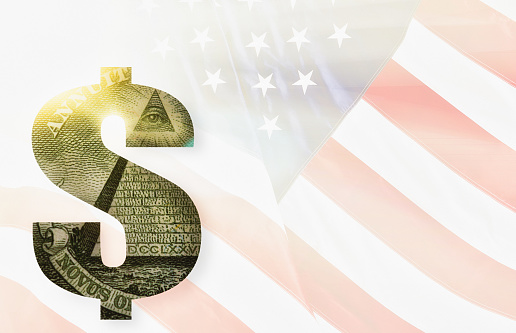 Glowing cut-out dollar currency symbol, made of a US $1 banknote, in front of the US national flag rippling gently in a breeze in the sunlight.