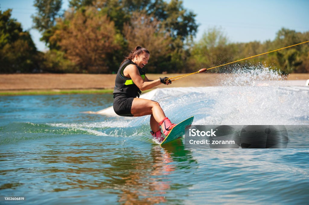 Smiling young woman tilting wakeboard upwards and spraying Happy young woman tilting wakeboard upwards and making a splash on water surface Wakeboarding Stock Photo