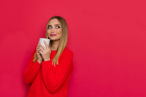 Cute young woman in red sweater is holding white cup, looking at copy space and smiling. Waist up studio shot on red background.